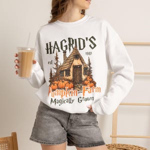 Può includere: Una felpa bianca a maniche lunghe con una stampa grafica raffigurante una capanna rustica circondata da pini e zucche arancioni. Il testo recita HAGRID'S est 1981 Pumpkin Farm Magically Grown in caratteri neri e arancioni dall'aspetto vissuto.