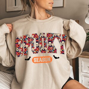 May include: Beige sweatshirt with the word "SPOOKY" in large, colorful letters filled with clown faces and flowers. Below the word "SPOOKY" is the word "SEASON" in an orange oval, with black bat silhouettes on either side.
