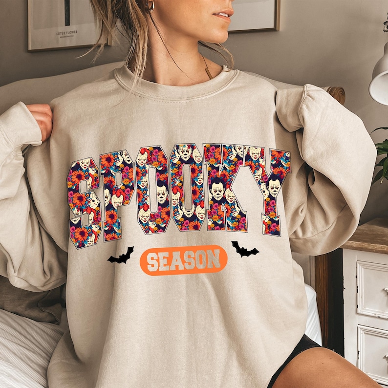 May include: Beige sweatshirt with the word "SPOOKY" in large, colorful letters filled with clown faces and flowers. Below the word "SPOOKY" is the word "SEASON" in an orange oval, with black bat silhouettes on either side.