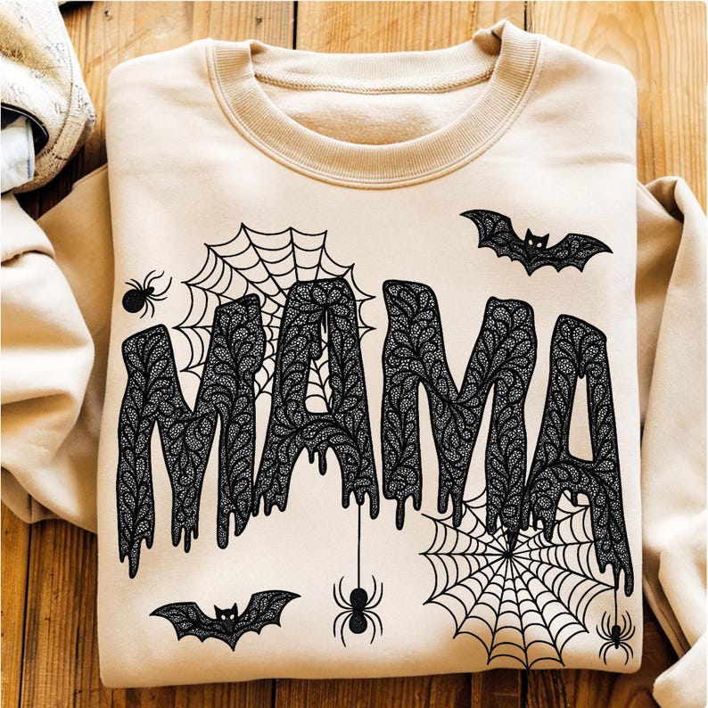 May include: Beige sweatshirt with the word "MAMA" in black, dripping, patterned letters. The design includes black spiders, spiderwebs, and bats, creating a Halloween theme. The sweatshirt is laid flat on a wooden surface.