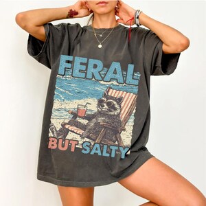 May include: A dark grey t-shirt featuring a graphic print of a raccoon wearing sunglasses and sitting in a beach chair with a drink. The text "FERAL BUT SALTY" is printed below the raccoon.