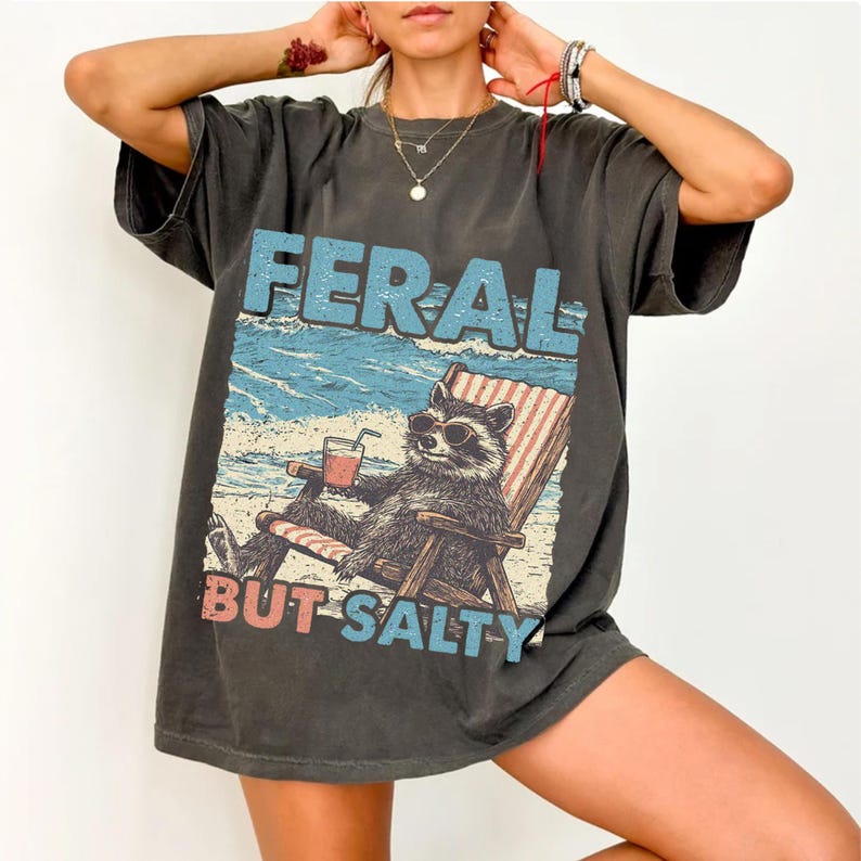 May include: A dark grey t-shirt featuring a graphic print of a raccoon wearing sunglasses and sitting in a beach chair with a drink. The text "FERAL BUT SALTY" is printed below the raccoon.