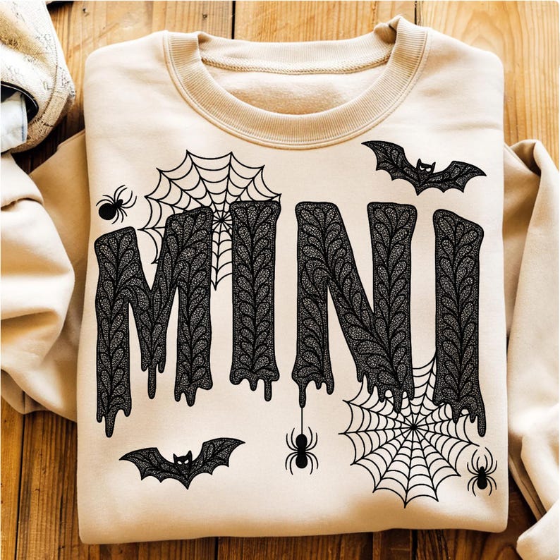 May include: A beige sweatshirt with the word "MINI" in large, black, dripping letters. The design includes spiderwebs, spiders, and bats, creating a Halloween theme. The sweatshirt is folded on a wooden surface.