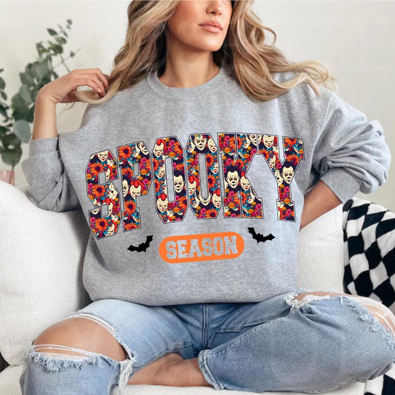 May include: A gray sweatshirt with the word "SPOOKY" in a floral design with clown faces. The word "SEASON" is below in an orange oval, with black bat graphics.