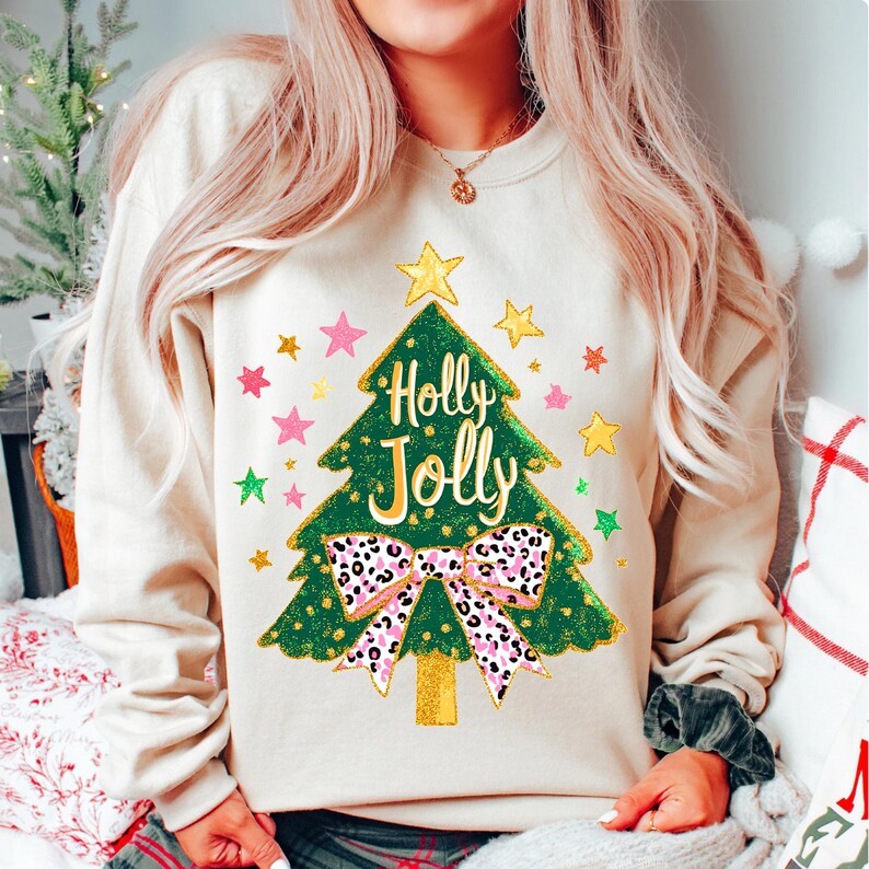 May include: Beige sweatshirt featuring a green Christmas tree design with the words "Holly Jolly". The tree has a gold glitter trunk and is decorated with pink, gold, and green stars. A pink and black leopard print bow is at the base of the tree.