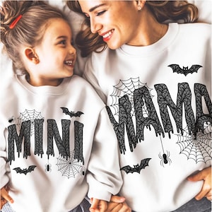 May include: White sweatshirts with black Halloween-themed designs. One sweatshirt says "MAMA" and the other says "MINI" in a dripping font. Both have spiderwebs, bats, and spiders. The image shows a mother and child.