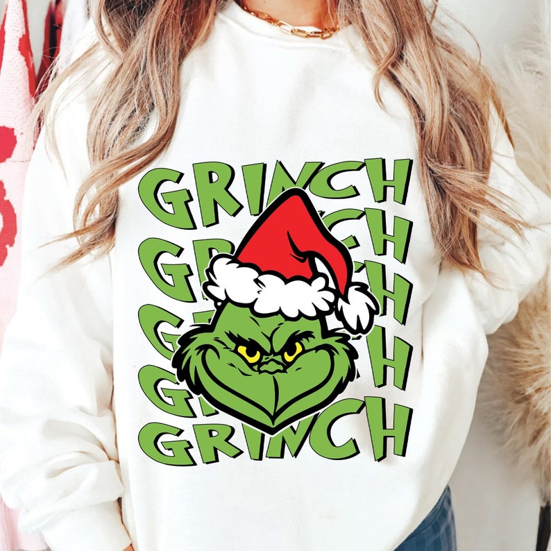 Puede incluir: Sudadera blanca con la cara verde del Grinch con un gorro de Pap&aacute; Noel rojo. La palabra "GRINCH" est&aacute; impresa en verde varias veces. Una prenda festiva con tem&aacute;tica navide&ntilde;a.