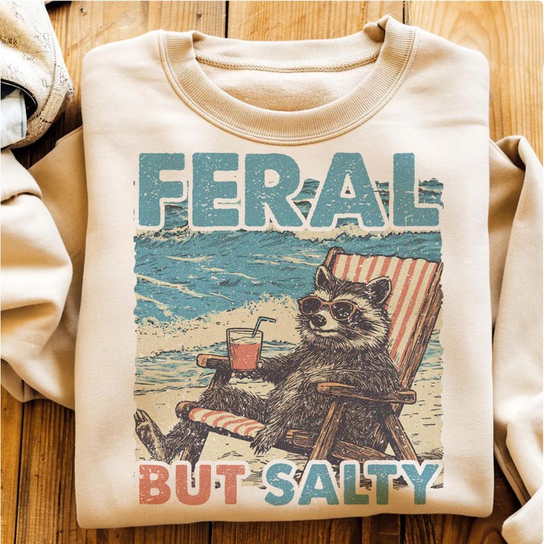 May include: A tan sweatshirt featuring a vintage-style graphic of a raccoon wearing sunglasses and relaxing in a beach chair with a drink. The text "FERAL BUT SALTY" is printed below the raccoon.