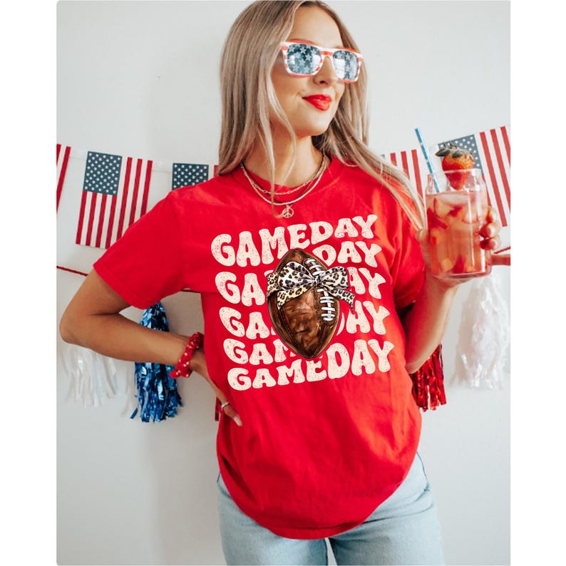May include: Red short-sleeved t-shirt with the text "GAMEDAY" repeated in a staggered pattern. A brown football with a leopard print bow is centred on the shirt.