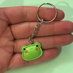 May include: A green frog keyring with a silver chain and ring. The frog has a stitched outline, black eyes, and orange cheeks. The keyring is held in a hand.