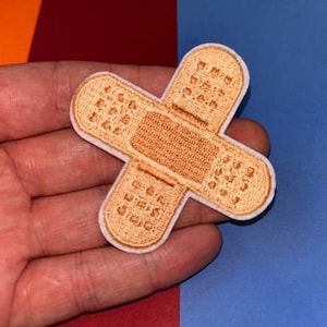 May include: Embroidered bandage-shaped patch, tan with a white border. The patch is in a cross shape, with a textured design. The background is a mix of red, orange, and blue.