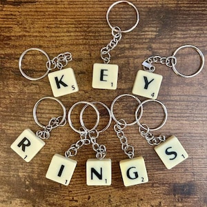 May include: A collection of keyrings spelling "KEY RINGS" using Scrabble tiles. Each keyring features a silver-coloured chain and ring. The tiles are off-white with black letters and numbers. The background is a wooden surface.