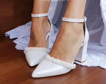 Puede incluir: Zapatos de novia de satén blanco con punta puntiaguda y tacón grueso. Los zapatos están decorados con una fila de perlas alrededor del tirante del tobillo y la punta.