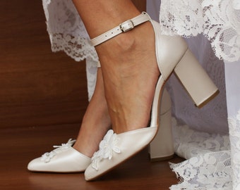 Puede incluir: Tacones de novia de color marfil con un tacón de bloque grueso y un delicado adorno floral en la punta.