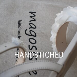 May include: White leather sandals with a white flower detail and the text "magos" and "HANDSTITCHED" on a beige fabric background.