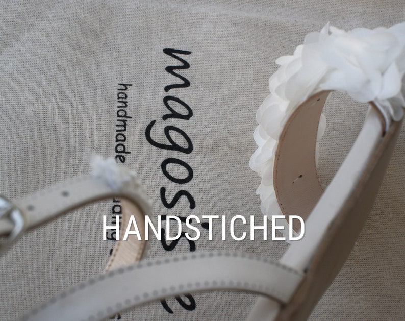May include: White leather sandals with a white flower detail and the text "magos" and "HANDSTITCHED" on a beige fabric background.
