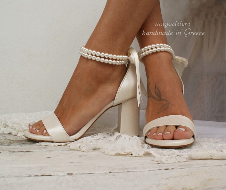 Puede incluir: Sandalias de tac&oacute;n blanco de sat&eacute;n con una correa de tobillo de perlas. Las sandalias tienen la punta cerrada y un tac&oacute;n grueso de bloque. El texto "magosisters handmade in Greece" es visible en la imagen.