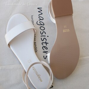 May include: A pair of white leather sandals with ankle straps. The sandals are handmade in Greece and have a brown leather sole. The text "magosisters" is printed on the straps.