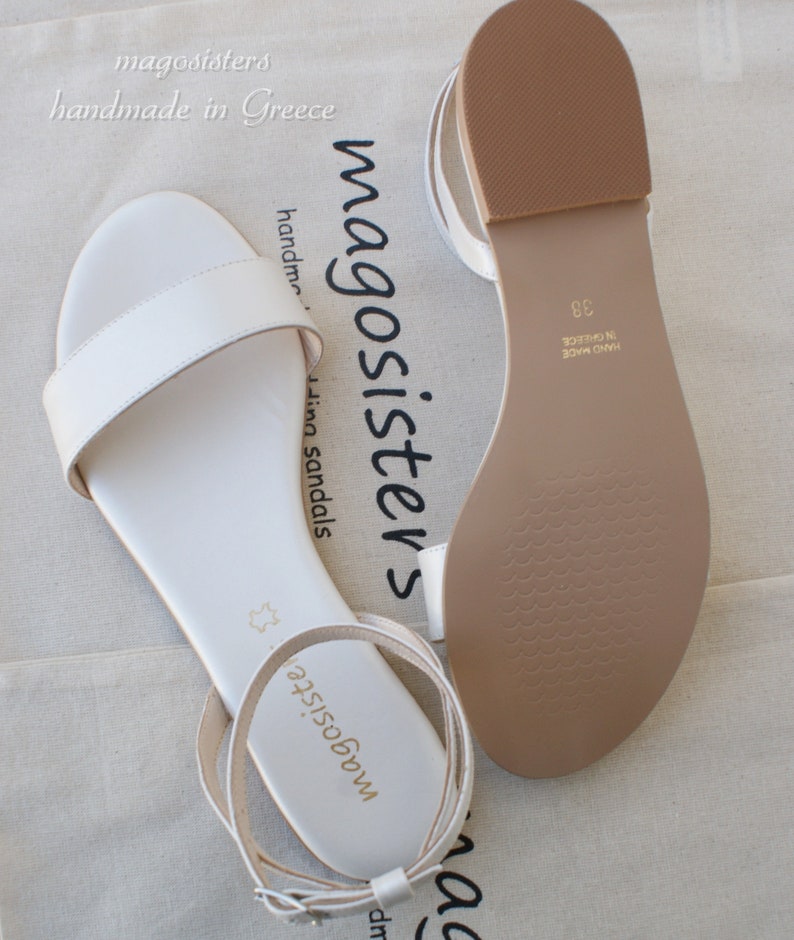 May include: A pair of white leather sandals with ankle straps. The sandals are handmade in Greece and have a brown leather sole. The text "magosisters" is printed on the straps.