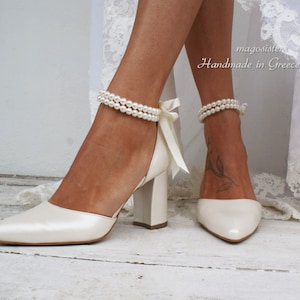 Op de afbeelding: Een paar ivoorwitte bruidsschoenen met parel enkelbandjes en satijnen strikken. De schoenen hebben een spitse neus en een blokhak. De schoenen staan op een wit houten oppervlak. "magosistem Handmade in Greece" staat op de achtergrond.