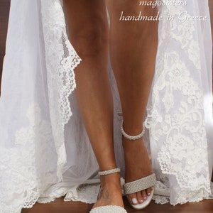 May include: White bridal sandals with pearl embellishments. The sandals have a delicate ankle strap and a low heel. The sandals are shown on a model wearing a white lace dress.