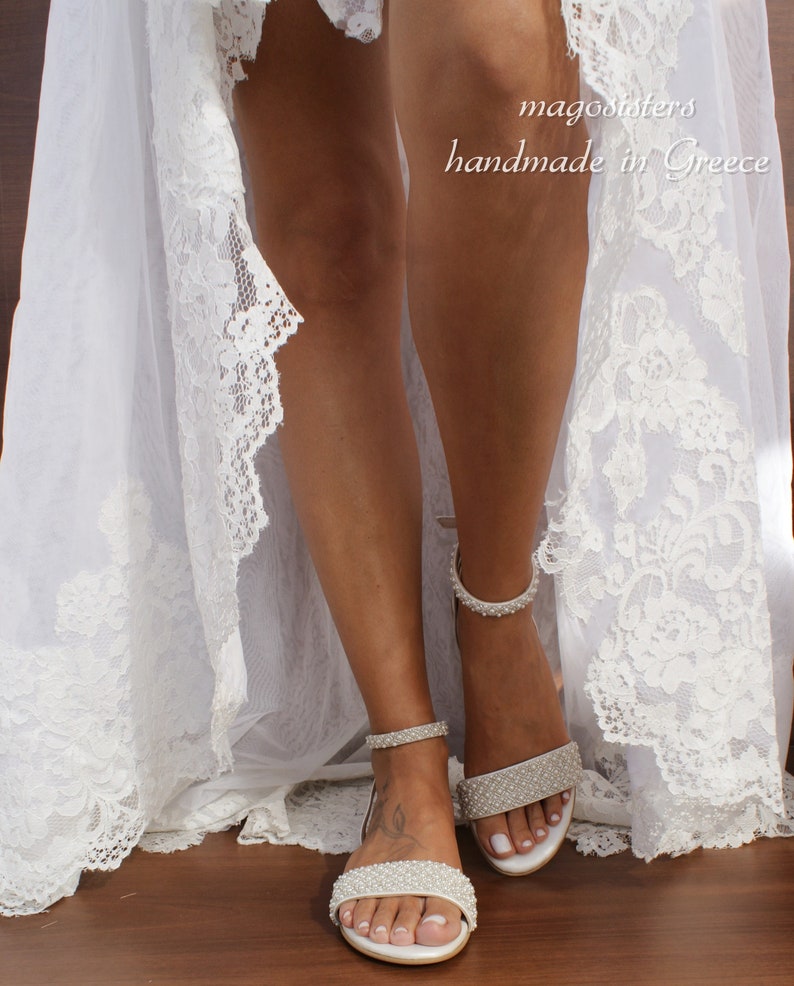May include: White bridal sandals with pearl embellishments. The sandals have a delicate ankle strap and a low heel. The sandals are shown on a model wearing a white lace dress.