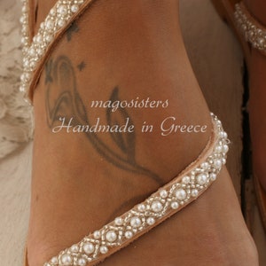 May include: A pair of white leather sandals with pearl embellishments. The sandals are handmade in Greece and feature a simple design with a single strap that wraps around the ankle.