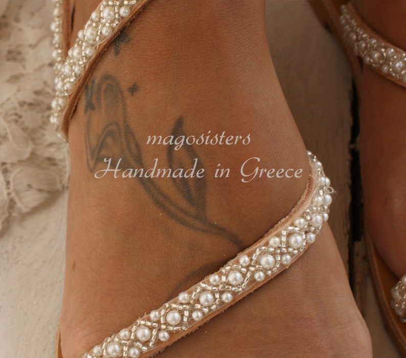 May include: A pair of white leather sandals with pearl embellishments. The sandals are handmade in Greece and feature a simple design with a single strap that wraps around the ankle.