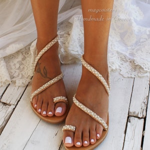 May include: Tan leather sandals with a pearl embellishment. The sandals have a toe ring and wrap around the ankle. The sandals are on a white wooden surface.  magoosisters Handmade in Greece
