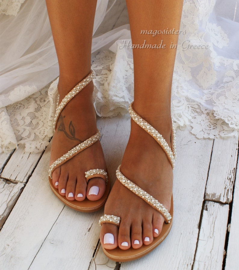May include: Tan leather sandals with a pearl embellishment. The sandals have a toe ring and wrap around the ankle. The sandals are on a white wooden surface.  magoosisters Handmade in Greece
