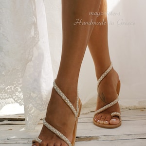 May include: A pair of tan leather sandals with pearl embellishments. The sandals have a toe ring and multiple straps that wrap around the ankle. The sandals are handmade in Greece.