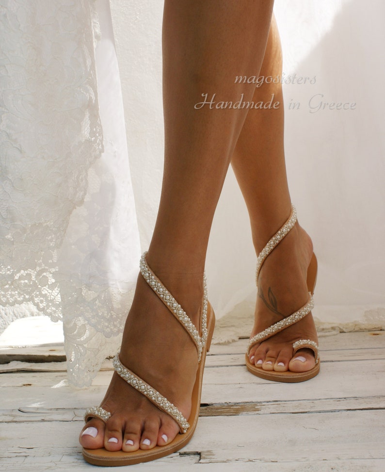 May include: A pair of tan leather sandals with pearl embellishments. The sandals have a toe ring and multiple straps that wrap around the ankle. The sandals are handmade in Greece.