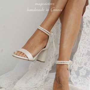Puede incluir: Sandalias de tac&oacute;n blanco de sat&eacute;n con una correa de tobillo de perlas. Las sandalias tienen un tac&oacute;n de bloque y una punta cerrada. El texto "magosisters handmade in Greece" es visible en la imagen.