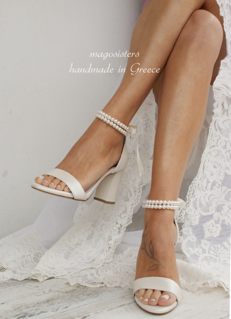Puede incluir: Sandalias de tac&oacute;n blanco de sat&eacute;n con una correa de tobillo de perlas. Las sandalias tienen un tac&oacute;n de bloque y una punta cerrada. El texto "magosisters handmade in Greece" es visible en la imagen.