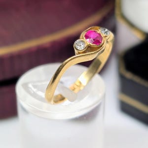 Könnte beinhalten: Ein Goldring mit einem zentralen, oval geschliffenen, leuchtend pink-roten Edelstein, der von zwei kleinen, rund geschliffenen, klaren weißen Steinen flankiert wird. Die Ringschiene hat eine glatte, polierte Oberfläche und eine geschwungene Form.