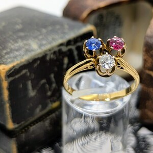 May include: A gold ring with three gemstones: a blue sapphire, a red ruby, and a white diamond. The ring is set in a delicate, floral design.