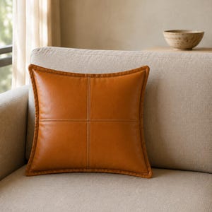 May include: A square throw pillow in a warm tan color, crafted from a smooth material. The pillow features a distinct cross-stitch pattern dividing the front surface into four equal quadrants, with piped edges framing the entire square shape.
