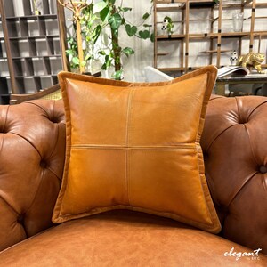 May include: A brown leather pillow with a square stitched design. The pillow is on a brown leather sofa.