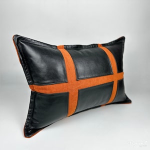 May include: A black leather cushion with orange suede accents. The cushion is rectangular and has a cross-shaped design.