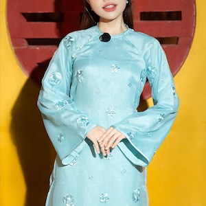 May include: A light blue dress with long sleeves and floral embroidery. The dress has a mandarin collar and a black pendant. The sleeves are flared at the wrist. The dress is made of a shiny fabric.