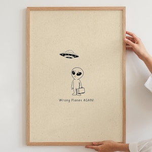 Puede incluir: Un póster enmarcado con un extraterrestre de dibujos animados y un OVNI. El extraterrestre lleva un maletín y mira hacia arriba. El texto "Wrong Planet AGAIN!" está debajo. El marco es de color marrón claro.