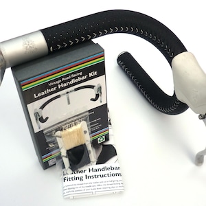 Puede incluir: Un kit de cinta de manillar de cuero negro con costuras blancas y una superficie perforada. El kit incluye una caja con instrucciones, un cepillo y una cinta de repuesto. La caja presenta un diseño de arcoíris y el texto "Vintage Road Racing Leather Handlebar Kit".