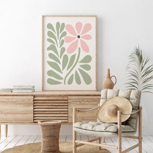 May include: A framed print with a pink flower and green leaves in a minimalist style. The print is hanging on a white wall above a wooden cabinet.