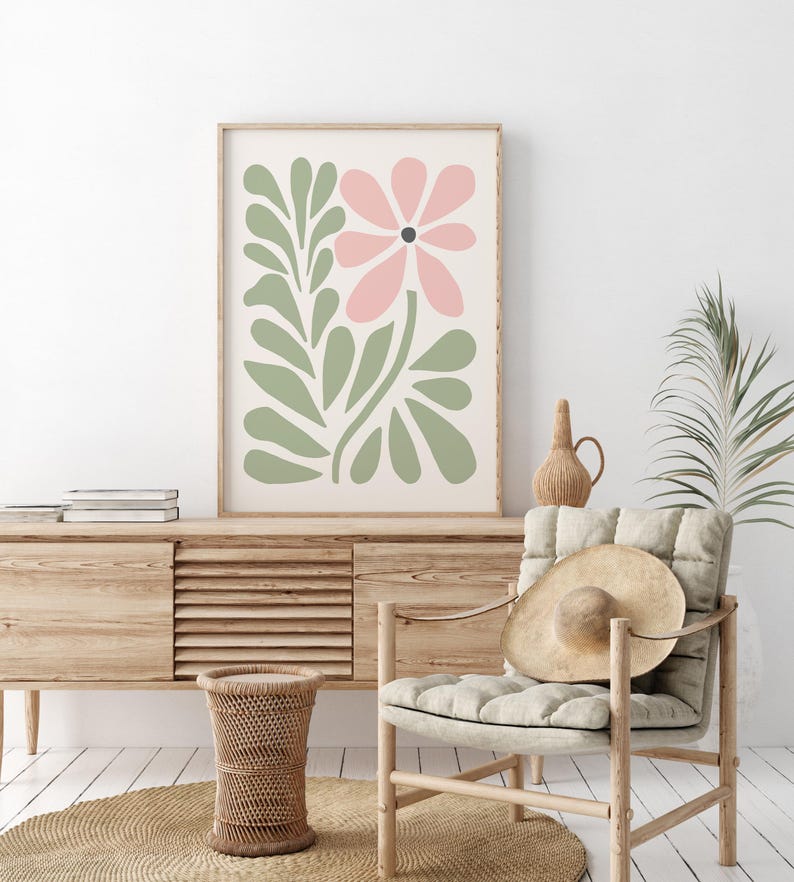 May include: A framed print with a pink flower and green leaves in a minimalist style. The print is hanging on a white wall above a wooden cabinet.