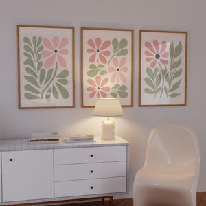 May include: Three framed prints with a floral design featuring pink and green flowers. The prints are hanging on a white wall above a white dresser with three drawers. A lamp is sitting on the dresser. A white chair is in the foreground.