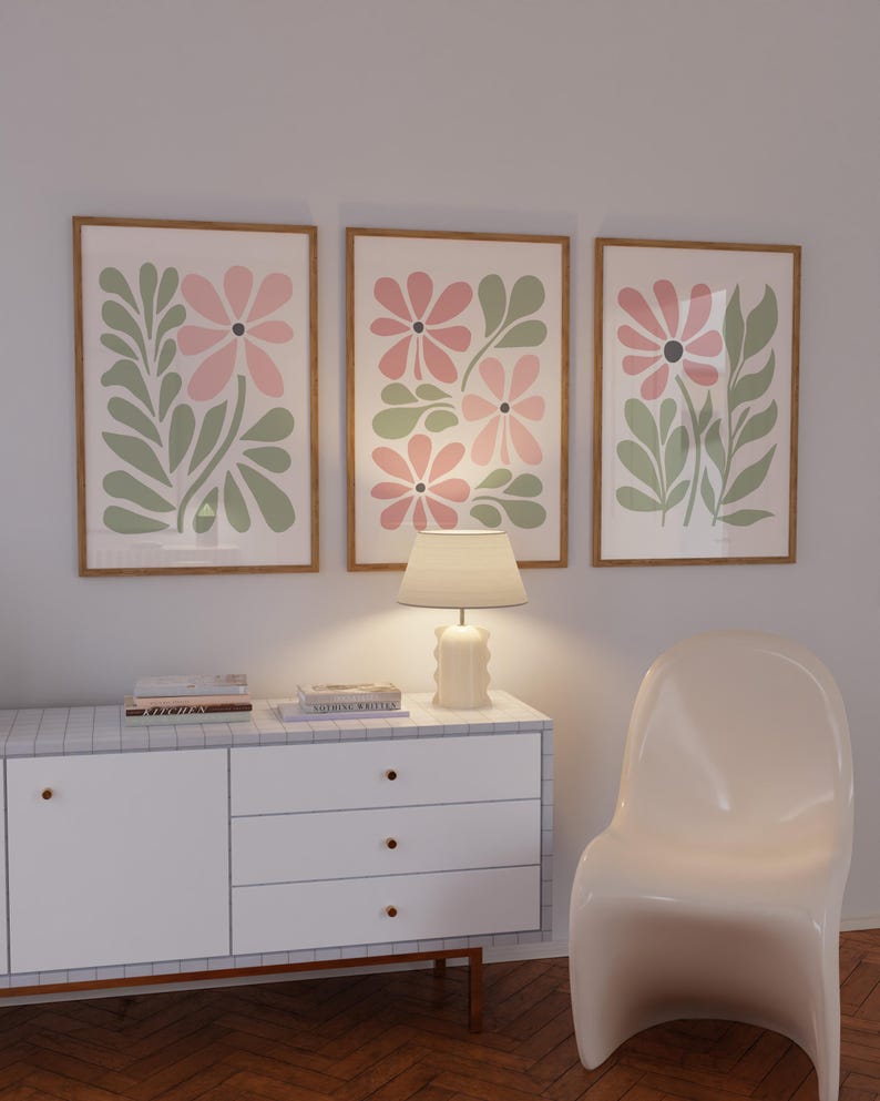 May include: Three framed prints with a floral design featuring pink and green flowers. The prints are hanging on a white wall above a white dresser with three drawers. A lamp is sitting on the dresser. A white chair is in the foreground.