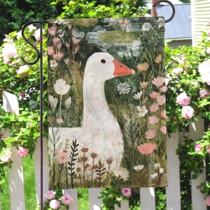 May include: A garden flag featuring a white goose with an orange beak, set against a backdrop of flowers and greenery. The artwork has a painterly style, with soft colors and a whimsical feel. The flag is designed for outdoor use and is displayed on a black metal stand.