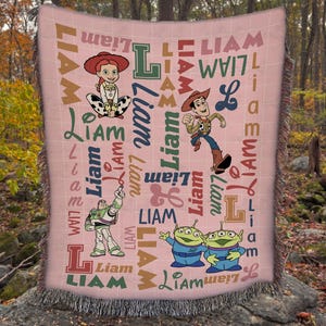 May include: A woven throw blanket with a light pink background featuring a repeating pattern of the name Liam in various fonts and colors, alongside illustrations of Jessie, Woody, Buzz Lightyear, and two green aliens from the Toy Story franchise.
