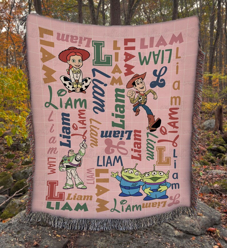 May include: A woven throw blanket with a light pink background featuring a repeating pattern of the name Liam in various fonts and colors, alongside illustrations of Jessie, Woody, Buzz Lightyear, and two green aliens from the Toy Story franchise.