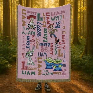 May include: A pink woven throw blanket featuring the name Liam repeated in various fonts and colors alongside illustrations of Jessie, Woody, Buzz Lightyear, and two green aliens from Toy Story. The blanket has a fringed edge on the top and bottom.